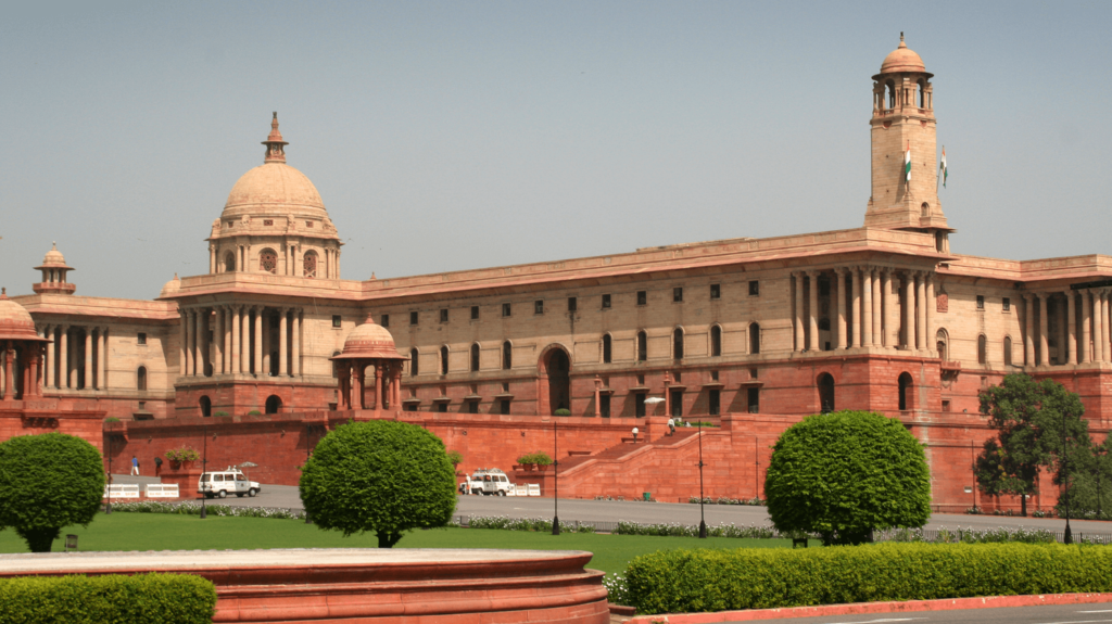The Supreme Court of India building on a sunny day, with well-maintained lawns and a clear sky.